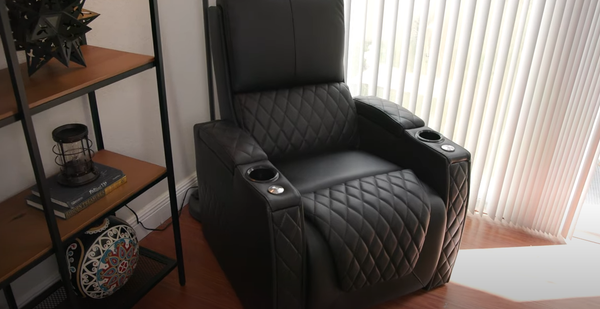 Black leather recliner chair with diamond stitch pattern, cup holders, and center console in a living room setting next to bookshelves and houseplant
